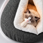 Brown-and-white cat resting inside a gray triangular cat cave bed with white plush lining, shown with Spanish on-screen captions.